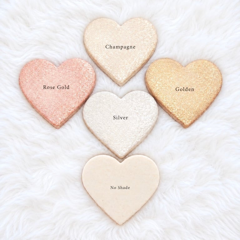 Five heart-shaped products arranged on white fluffy fabric, labeled Champagne, Rose Gold, Golden, Silver, and No Shade