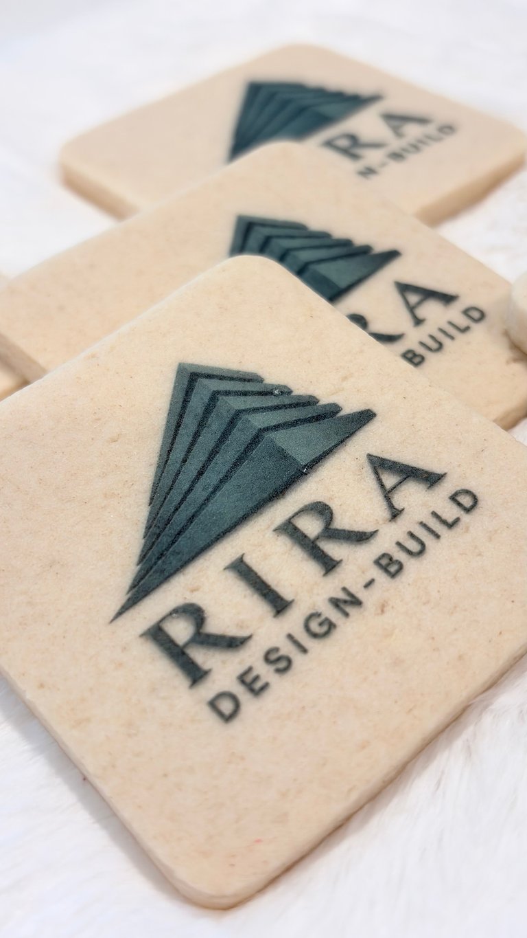 Three cream-colored wooden coasters with RTRA Design Build logo featuring a geometric arrow design in dark teal ink